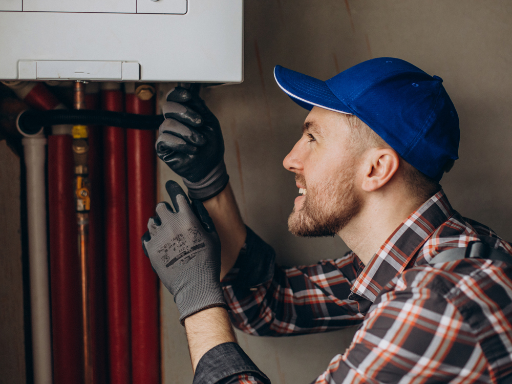 A man wearing a blue hat and plaid shirt repairs a water heater, demonstrating his skills in home maintenance.